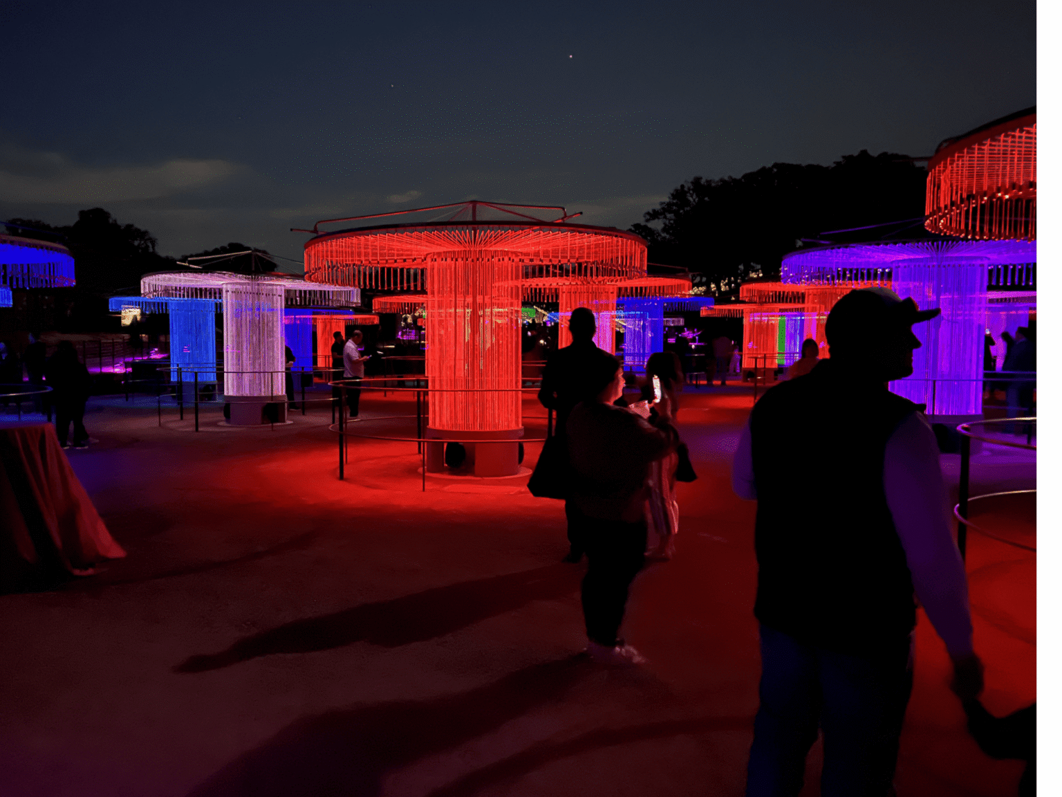 Numerous visitors walk among brightly lit, cylindrical art installations at night, casting colorful reflections on the ground.