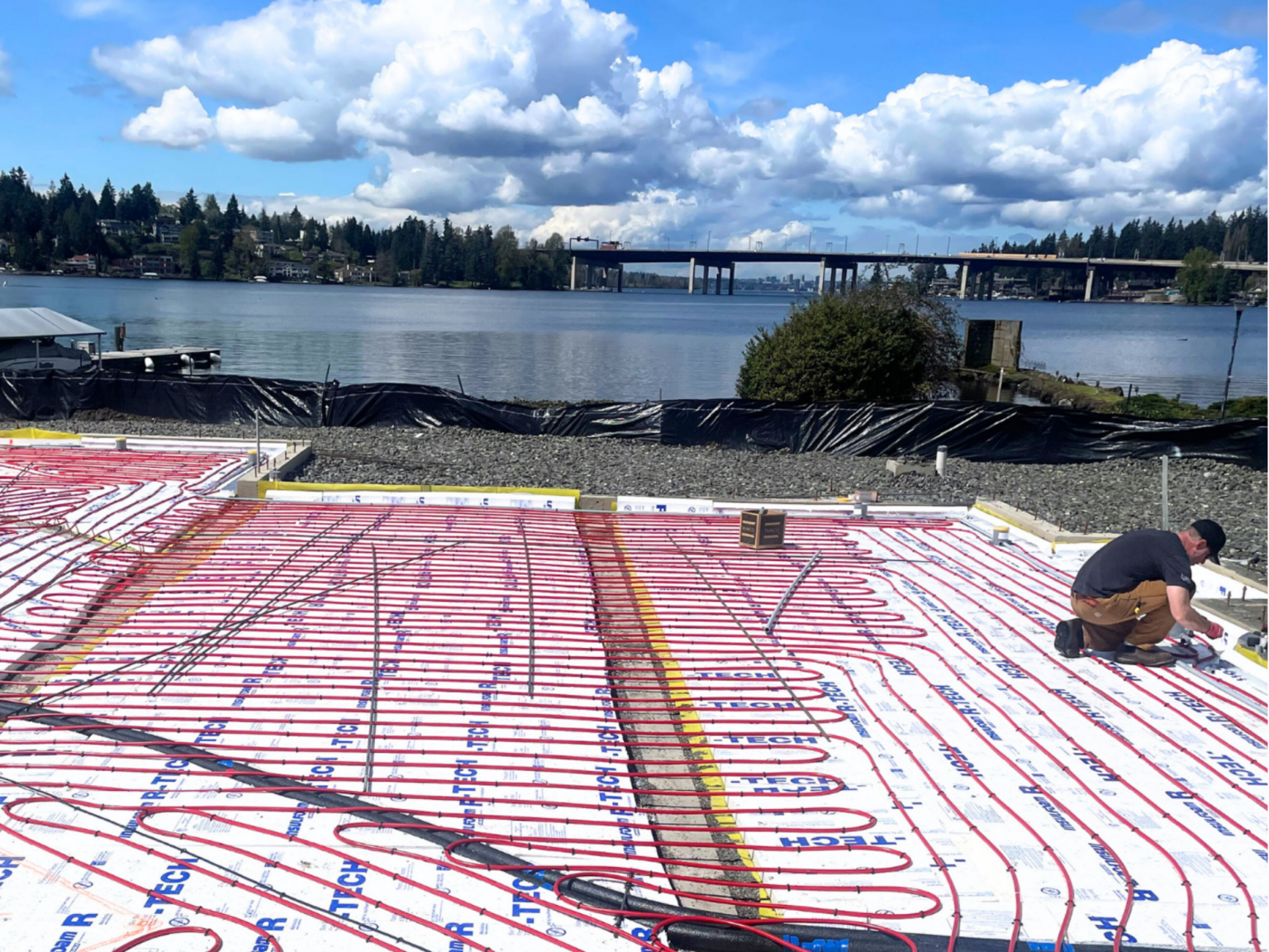 Radiant floor heating system, red PEX tubes, installed on concrete. A worker kneels by a lake, a bridge in the distance.