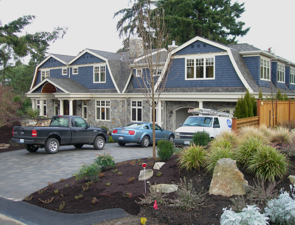 Spacious blue and stone home featuring a prominent garage. A pickup, sports car, and work van sit on the driveway amidst new garden beds.