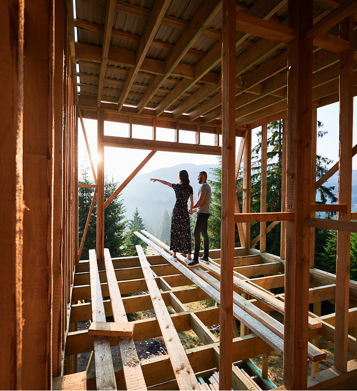 A couple holds hands inside an unfinished wood-framed house, looking out at a sunlit forest and mountain landscape.