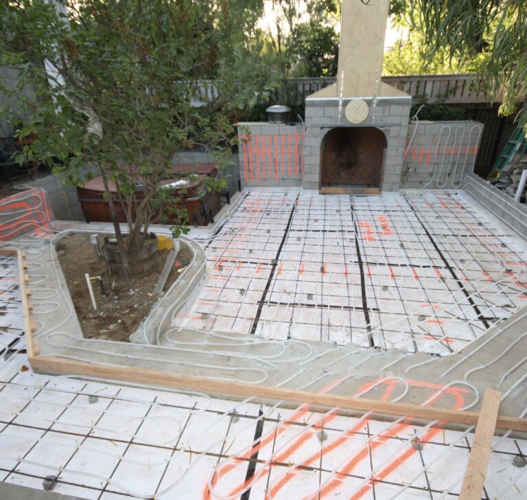 Backyard patio under construction with orange radiant heating PEX tubing on a rebar grid. A stone fireplace and hot tub are nearby.