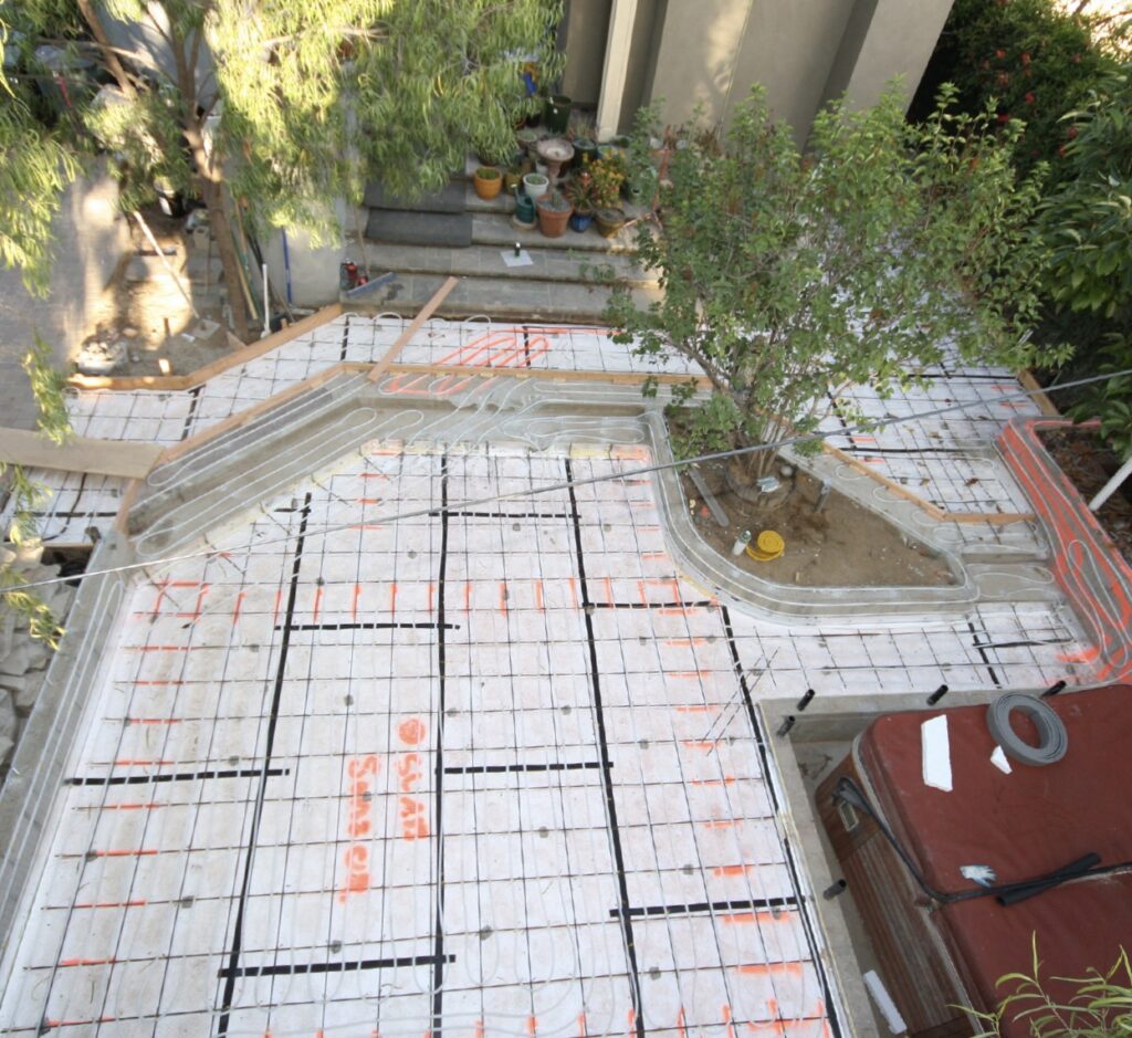 Backyard patio under construction with orange radiant heating PEX tubing on a rebar grid. A stone fireplace and hot tub are nearby.