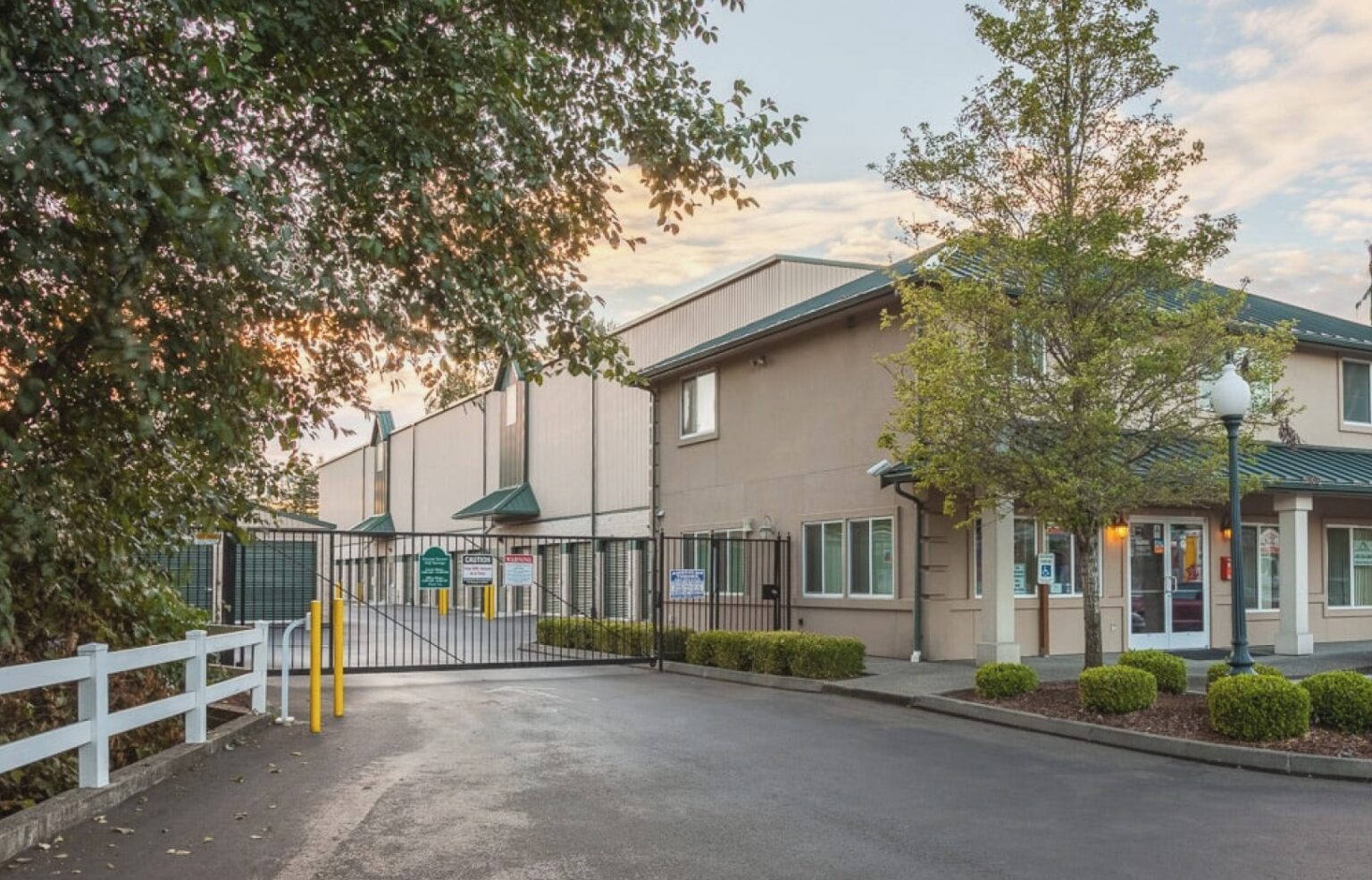 Secure self-storage facility entrance with a closed metal gate, a multi-story office building, and a large storage unit building beyond.