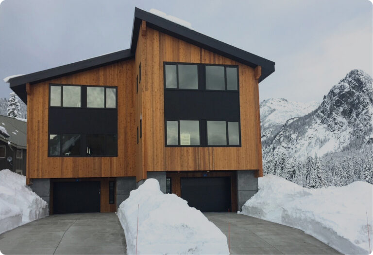 Contemporary wood and dark-paneled home, featuring two garage doors, surrounded by heavy snowdrifts and dramatic snowy peaks. The driveway and walkways are clear with mounds of snow on either side.