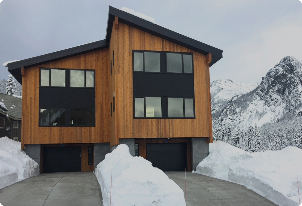 Contemporary wood and dark-paneled home, featuring two garage doors, surrounded by heavy snowdrifts and dramatic snowy peaks. The driveway and walkways are clear with mounds of snow on either side.