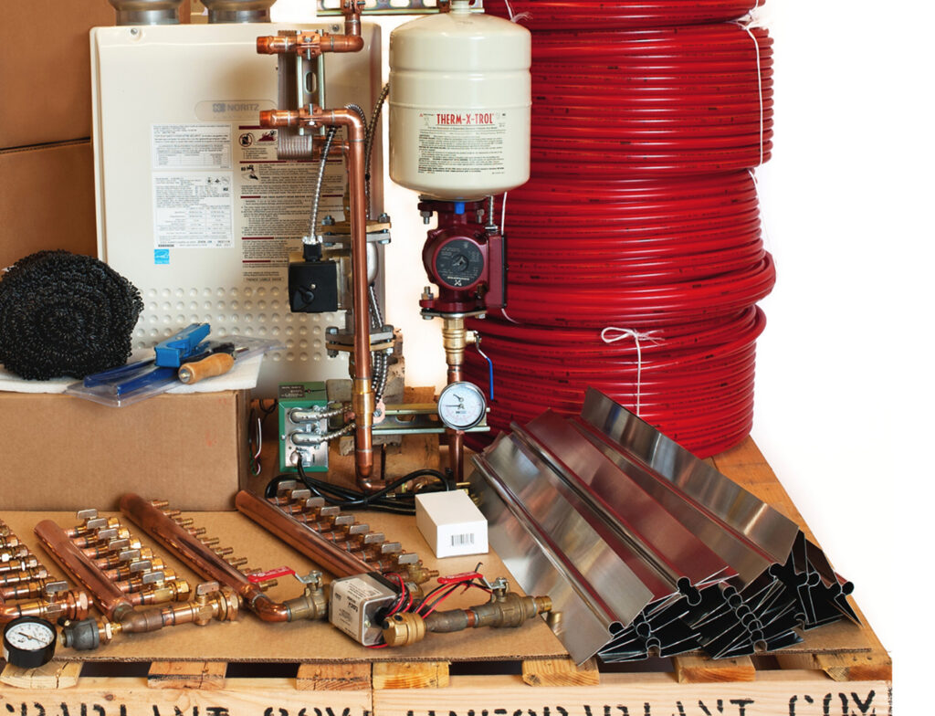 Radiant heating system supplies: white boiler, copper pipes, red PEX tubing rolls, and metal heat transfer plates on a wooden pallet.